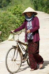 Vietnam  Frau schiebt ihr Fahrrad ueber eine Landstrasse