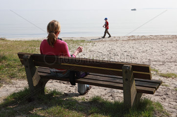 Klink  Deutschland  Frau sitzt auf einer Bank am Strand und beobachtet ihr Kind