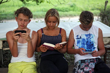 Bolsena  Italien  Jungen spielen mit ihren Smartphones waehrend ein Maedchen in einem Buch liest