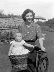Woman on a bicycle with a baby riding in a basket  c 1955