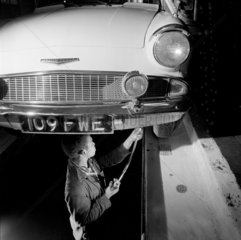 A young black garage fitter in pit greases a Ford Anglia car  1964.