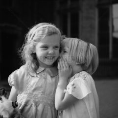 Small girl whispering into her friend’s ear  c.1930s.