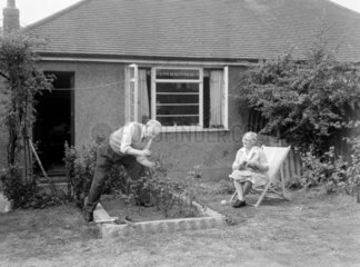 Man gardening as a woman watches from a deckchair  1955.