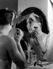 Man shaving using a razor in a mirror  c 1950s.