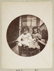 Two children sitting in a wicker chair outside their house  c 1890s.