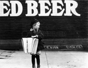 Young newsboy in front of a saloon  USA  1910.