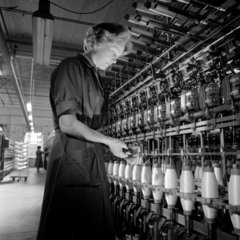 A female mill worker winds glass based fabrics onto bobbins in twisting room.