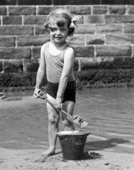 Small girl shovelling wet sand into a bucket on the beach  c 1930s.