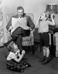 Two children playing while a man reads a newspaper  c 1949.