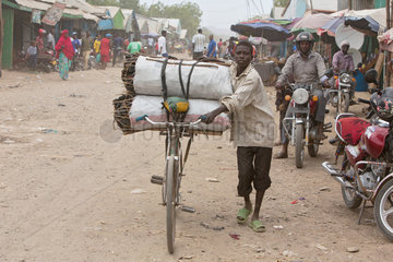 Kakuma  Kenia - Strassenszene. Ein Junge transportiert Holzkohlesaecke auf seinem Fahrrad auf einer belebten unbefestigten Strasse.