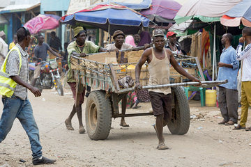 Kakuma  Kenia - Tageloehner ziehen einen grossen Transportanhaenger  beladen mit Waren auf einer belebten  unbefestigten Strasse.