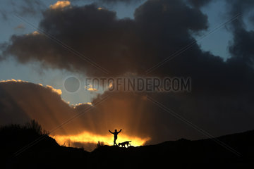 Wustrow  Deutschland - Silhouette  Junge laeuft bei Sonnenuntergang mit seinem Hund ueber eine Duene