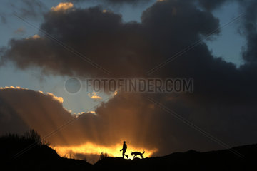 Wustrow  Deutschland - Silhouette  Junge laeuft bei Sonnenuntergang mit seinem Hund ueber eine Duene