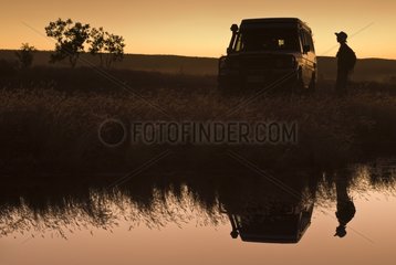 Reflections of adventurer woman and car in a pond at sunset