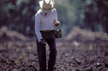 Mexico; a farmer wearing a cowboyhat while sowing corn from a small traditional sowing seed bag into the bare soil