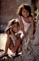 Mexico  Mexico City; two youngh girls smiling while leaning against a tree trunk
