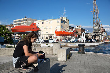 Kroatien  Rijeka - Maedchen sitzt am Hafen  beschaeftigt mit smartphone und social media