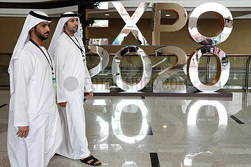 Dubai  Vereinigte Arabische Emirate  Maenner in Landestracht laufen an einem Werbeaufsteller fuer die Expo 2020 vorbei