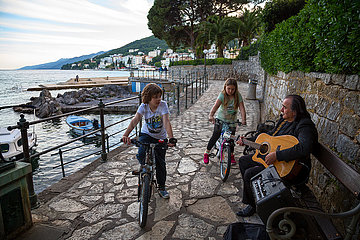 Kroatien  Opatija - Mann mit Gitarre singt Balladen  zwei Kinder auf Fahrraedern hoeren ihm zu