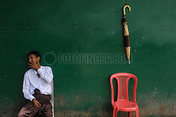 Yangon  Myanmar  Mann sitzt in der ehemaligen Hauptstadt neben einem roten Stuhl und einem Regenschirm