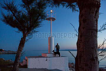 Kroatien  Krk (Hauptstadt der Insel Krk) - Mann mit Hund am Leuchtturm des Yachthafens in der Abenddaemmerung