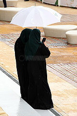 Riad  Saudi-Arabien  Arabische Frauen stehen bei Regen unter einem Regenschirm und schauen auf ein Smartphone