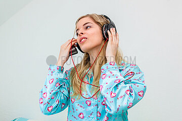 junge Frau im Schlafanzug sitzt auf ihrem Bett und hört Musik mit Kopfhörer