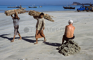 Thandwe  Myanmar  Einheimische tragen Brennholz und gehen am Strand von Ngapali Beach an einem Touristen vorbei