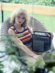 Berlin  Deutsche Demokratische Republik  junge Frau sitzt in einem Korbsessel und hoert Musik aus einem Kofferradio