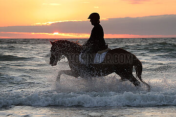 Dierhagen  Silhouette: Frau reitet auf ihrem Pferd bei Daemmerung durch das Wasser der Ostsee